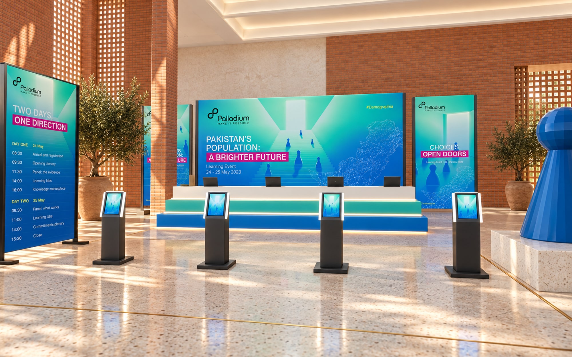 A registration desk built as three steps in white, teal and blue with light under each, four check-in kiosks in front of it, the key visual and two banners on frames behind, an agenda board at the left