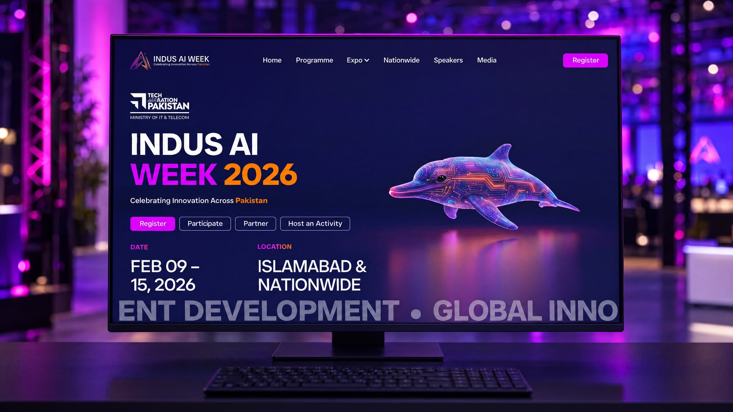 The Indus AI Week homepage on a large desktop display in the press area of a technology summit, the hall behind it in magenta and violet bokeh