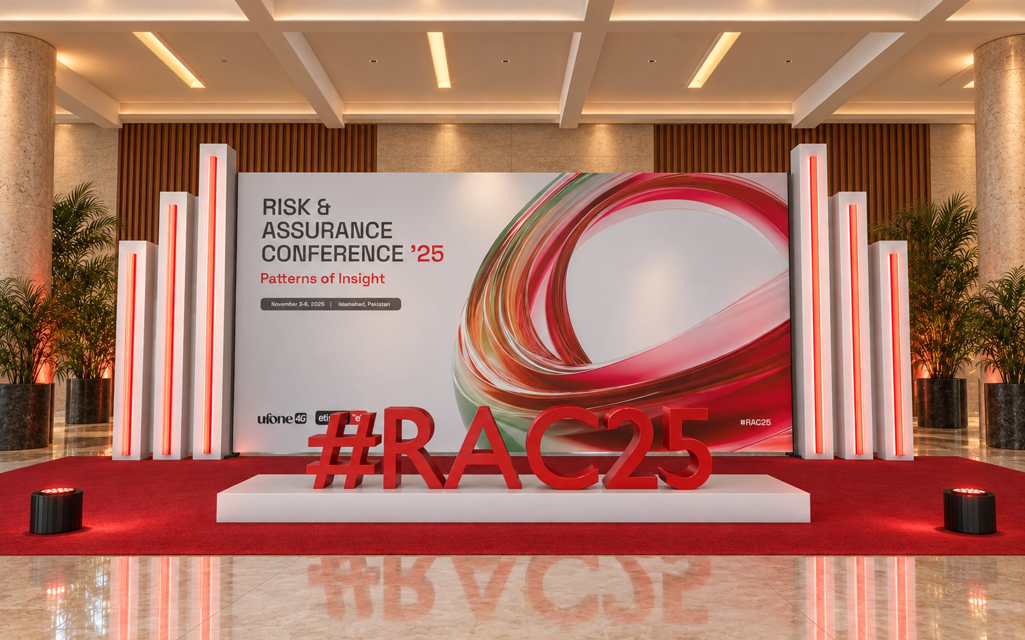 A media wall carrying the key visual between stepped white fins of red light, with large red three-dimensional letters reading #RAC25 on a white plinth in front of it