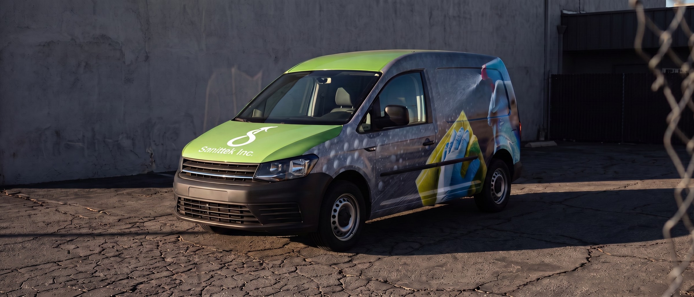 A wrapped Sanittek van parked on cracked asphalt in low late sun, green cab and a full-bleed side graphic.