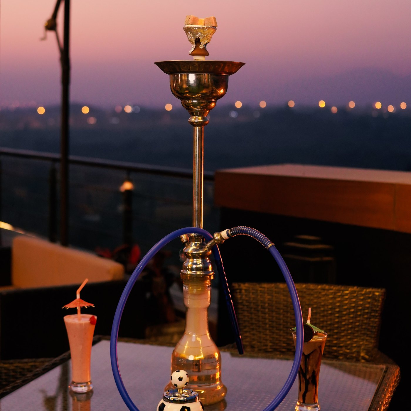 A shisha pipe and two drinks on a rooftop terrace table at dusk, city lights behind.