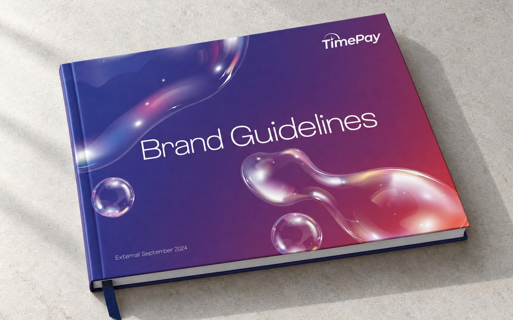 The brand guidelines book closed on pale stone, its gradient cover with the bubbles and the words Brand Guidelines, a navy ribbon marker