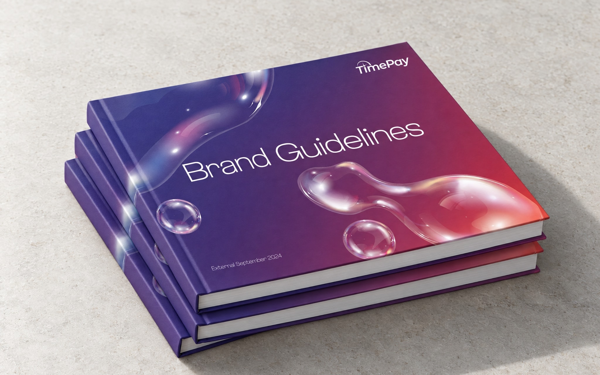 Three copies of the brand guidelines book stacked and fanned on pale stone, the top cover showing the gradient and the title