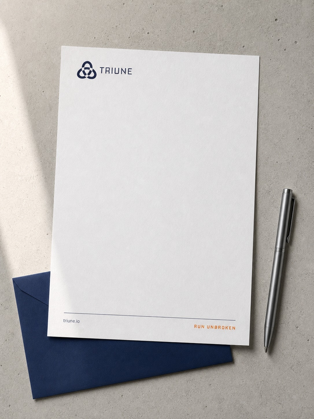 An A4 Triune letterhead shot from above on a paper surface, lockup at the top left, tagline in orange at the foot, with a navy envelope and a pen beside it