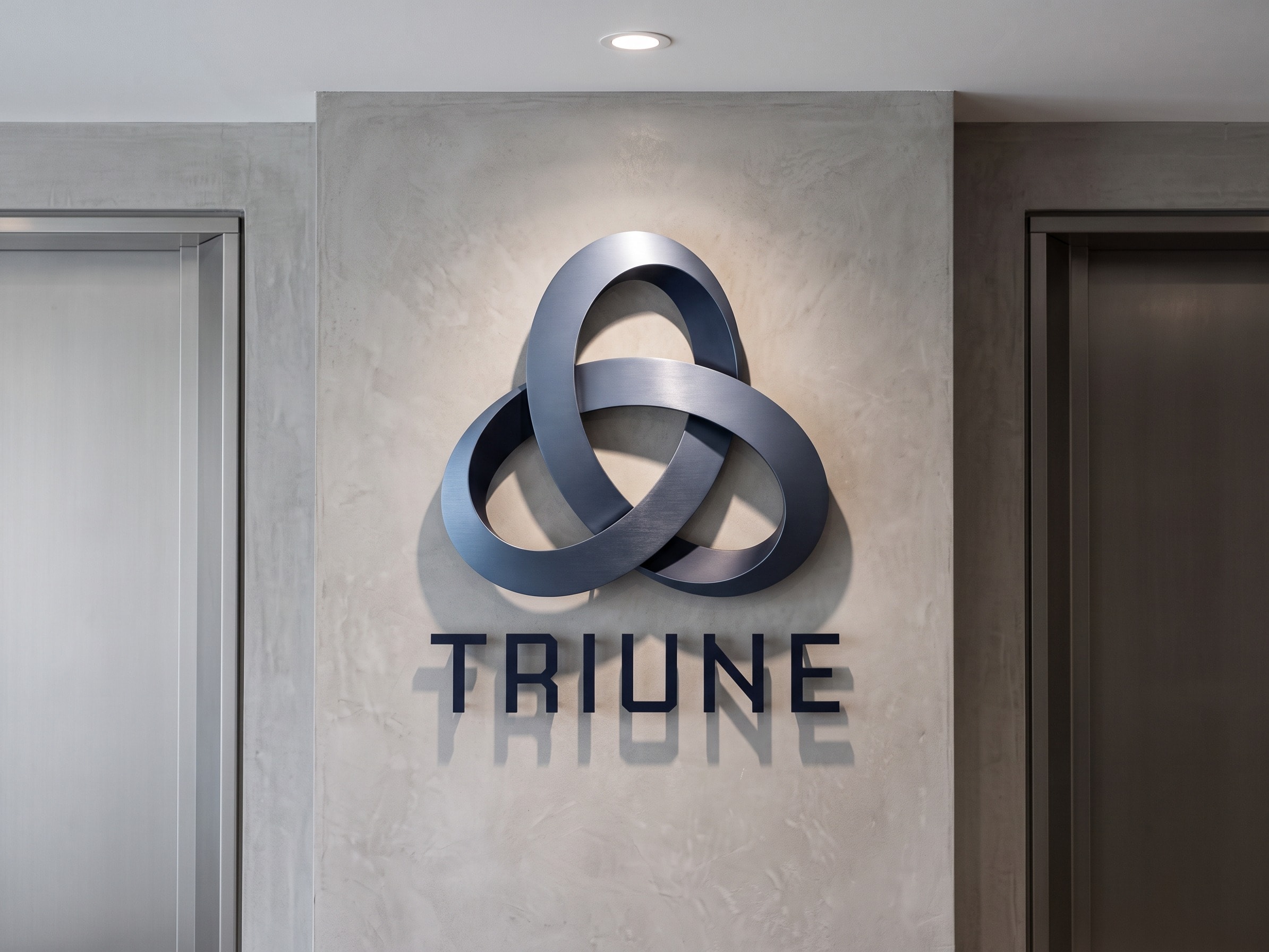 The stacked Triune lockup on a pale microcement pier between two lift doors, the symbol machined in navy and the wordmark cut flat below it