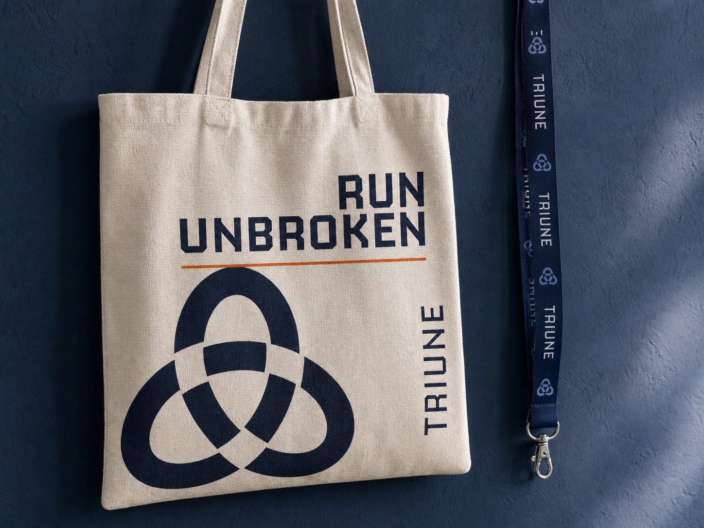 A natural canvas tote printed in navy: the tagline set large across the top, the mark cropped off the bottom edge, the wordmark running up the side seam, with a patterned lanyard beside it